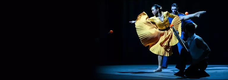 A dancer in a yellow dress strikes a one-legged pose with her remaining arms and legs extended. Another dancer is running towards her from behind. 