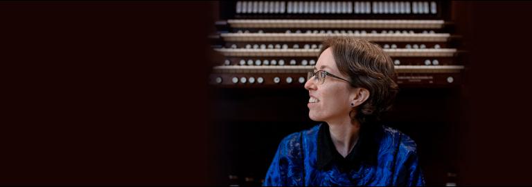 An organist wearing a deep blue button down with short, dark brown hair and glasses sits on a bench in front of a large, wooden organ console with their head turned to the right. 