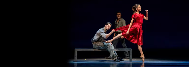 Dancer in red dress in arabesque, another pulls the hem of the dress. He's wearing dirty work clothes and sitting on a bench.