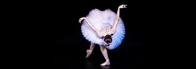 A ballerina wearing a white tutu, crown and pointe shoes, hits a low curtsy position, with their upper back facing the audience as they bend forward