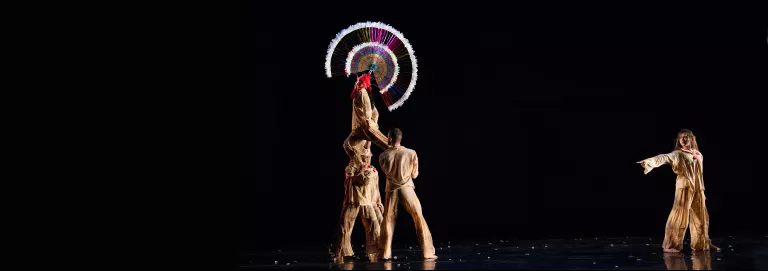 Two dancers hold a third above their shoulders, while a fourth dancers across the stage points a finger at the elevated dancer. The elevated dancer wears a large, circular headpiece, that stands out before the all black background.
