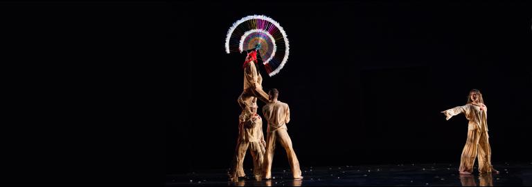 Two dancers hold a third above their shoulders, while a fourth dancers across the stage points a finger at the elevated dancer. The elevated dancer wears a large, circular headpiece, that stands out before the all black background. 