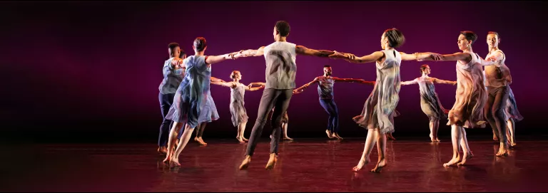 A group of dancers formed in a circle all holding each other by the wrists. The stage is lit with an ombre of black, purple and pink.