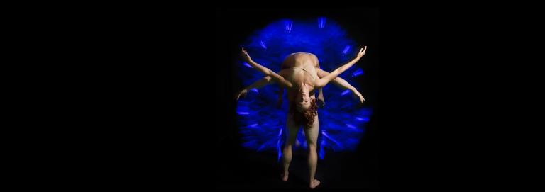 A dancer stands with their back towards the camera. Another dancer in a nude bra and underwear hangs from the shoulders of the standing dancer, in a backbend position with their chest and face towards the audience.