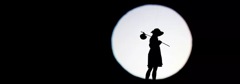 A black rectangle with a white spolight, reveals the figurine/shadow of a dancer wearing a dress, bonnett, and caryying a bindle over their shoulder.