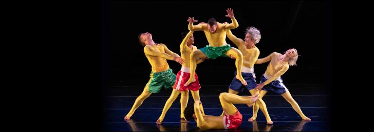In Front of a black background, six dancers wearing yellow bodysuits and blue, green, and red shorts hold each other in various positions. 