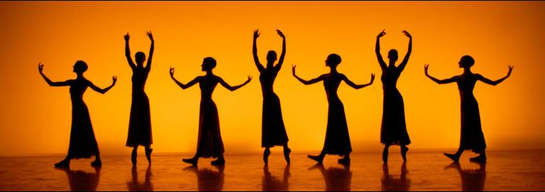 A stage is lit with a golden yellow hue, revelaing the shadows of seven dancers, four of which hold thier arms in a position parallel with the floor, three of which hold thier arms above their heads. 