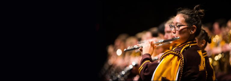 A UMN marching band member dressed in maroon and gold, playing the flute on stage. The camera focus is on the one player, while rows of other players are blurred in the background. 