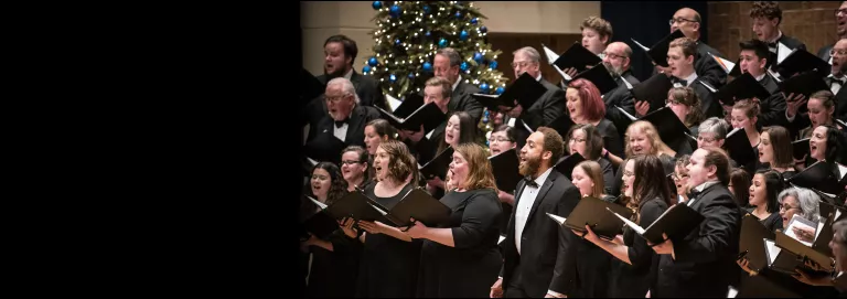 A large group of varying genders and ages all dressed in black and white, stand on elevated rows, singing in choir, with a green, lit Christmas tree in the background.