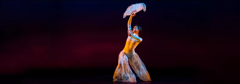 A dancer poses onstage, looking off into the distance with one hand outstretched toward the camera and the other upwards, holding a handheld fan. 