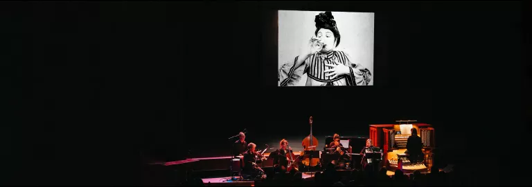 An ensemble of 7 musicians dressed in black perform onstage with a projection of a film above them. The screen shows a black and white film still of a person wearing a striped top, drinking a glass of wine with one hand held to their chest and their eyebrows raised.