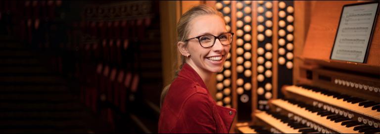 Anna Lapwood with blond hair and black glasses dressed in a red jacket smiles, sitting in front of a pipe organ with their head turned towards the camera.