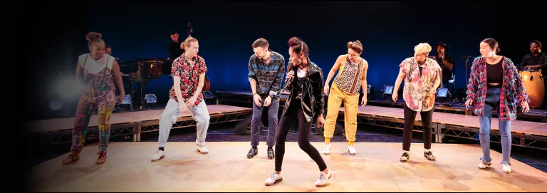 Six dancers stand on stage wearing casual clothing and tap shoes. One dancer is slightly downstage while the other five dancers form a line behind them.