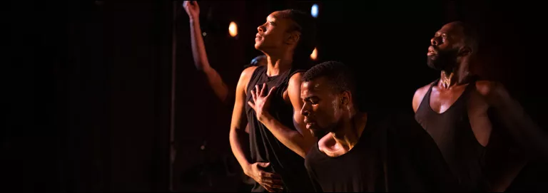 Three dancers stand onstage in a group wearing black, loose-fitted clothing. One dancer is slightly upstage and looks down while the other two look up and to the right.