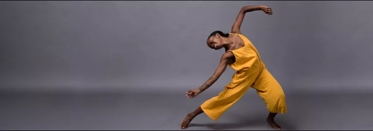 A dancer wearing a yellow, sleeveless jumpsuit poses against a gray backdrop. They face the camera with their head bent to the left and parallel to the ground with one leg stretched out to the left and the other bent slightly at the knee. One arm drapes across the outstretched leg, the other raised up and bent at the elbow.