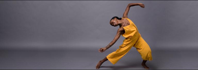 A dancer wearing a yellow, sleeveless jumpsuit poses against a gray backdrop. They face the camera with their head bent to the left and parallel to the ground with one leg stretched out to the left and the other bent slightly at the knee. One arm drapes across the outstretched leg, the other raised up and bent at the elbow.