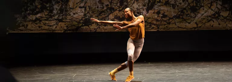 A dancer performs onstage dressed in a yellow top and shoes, and white pants. They stretch their arms out to the left and across their body, which faces the camera. One leg stands in front of the other, slightly bent with the other crossed behind.