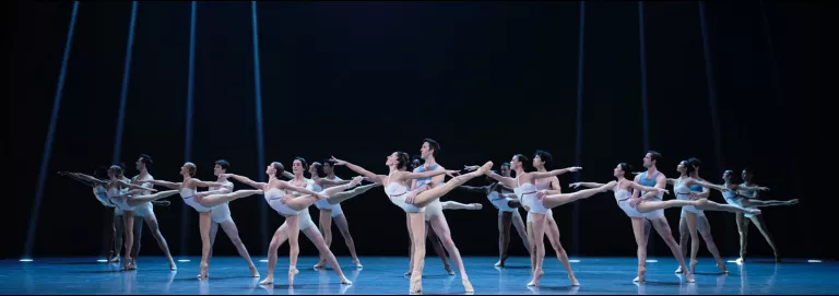 A group of dancers in white costumes are partnered up, and looking to the left. Front dancers are on pointe in arabesque, while their partner hold their waists.