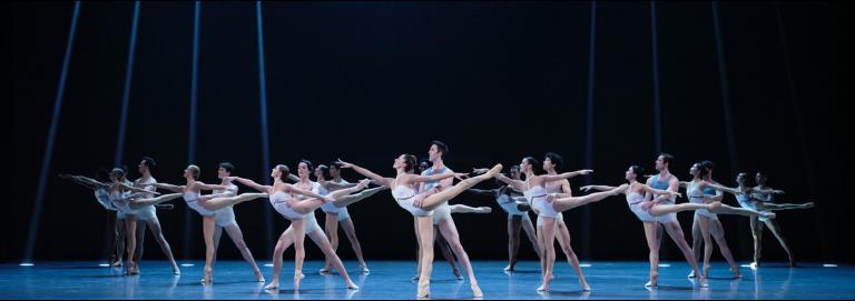 A group of dancers in white costumes are partnered up, and looking to the left. Front dancers are on pointe in arabesque, while their partner hold their waists.
