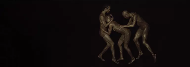 Three dancers appear in dark lighting. Two dancers embrace while the other follows closely behind with their hands placed on one of the dancer's back.