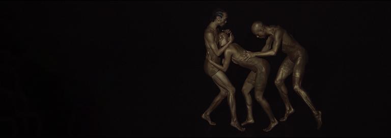 Three dancers appear in dark lighting. Two dancers embrace while the other follows closely behind with their hands placed on one of the dancer's back.