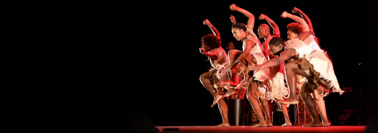 A group of dancers stand closely together onstage dressed in white clothing with their right arms and left legs raised, gazing downward toward the left and accompanied by a soft glow of red lighting.
