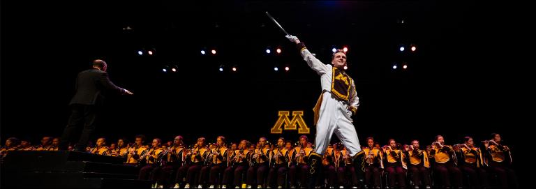 A drum major wearing white, maroon, and gold stands downstage with a large marching band grouped behind them. A University of Minnesota letter "M" logo appears in the background.