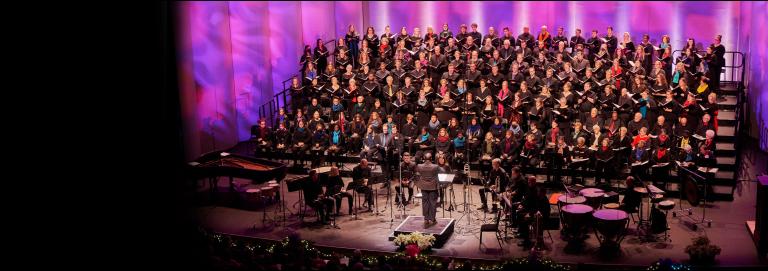 A choir wearing black with various colored scarves performs onstage with patterned pink and purple lighting projected behind them. A conductor stands downstage with their back toward the camera and musicians surrounding them.
