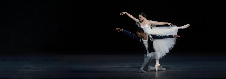 Ballerina hovers behind male parter who is on one knee. Their arms outstretched alike.