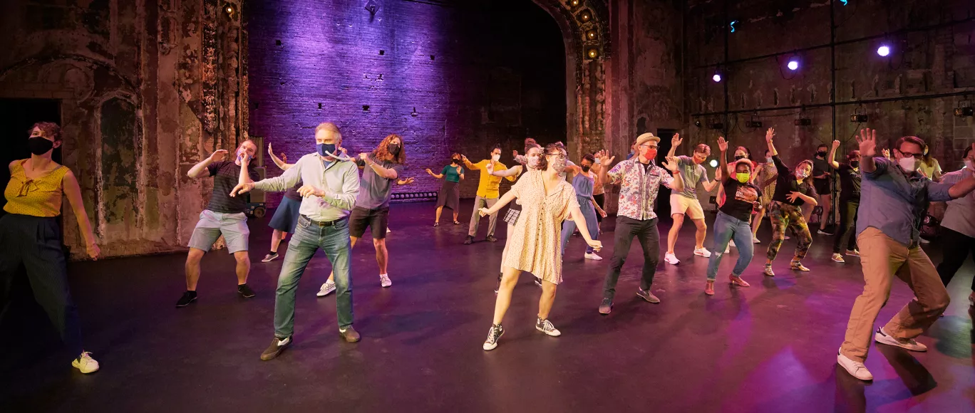 A large group of dancers participating in a dance class, stand on a stage that is lit with purple lights. All dancers are wearing masks while striking a standing pose with both arms out to either side. 