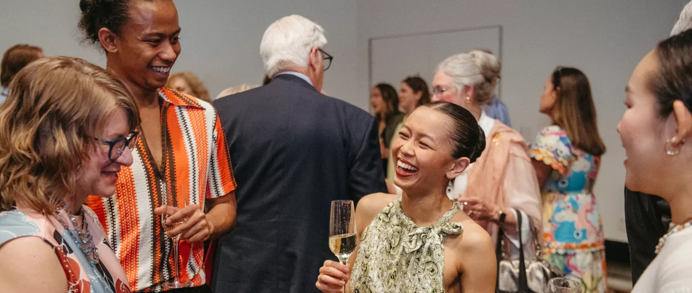 Four people engage in a cheery conversation at a cocktail party. From left to right there is a women with short blonde hair, glasses, and a blue blouse, a man in an orange button down shirt holding a champagne glass, a women in a slicked-back bun and green tank top holding a champagne glass, and another women with slick black hair and a white blouse.