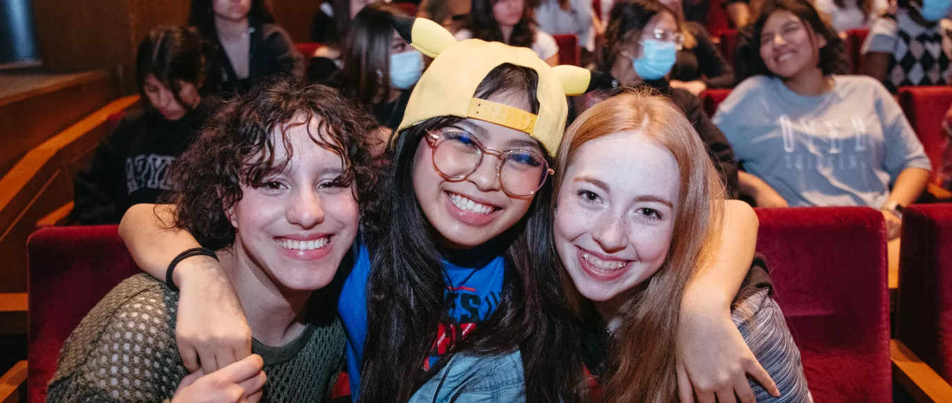 Three students sit in the Carlson Family Stage seats, smiling. The person in the middle has their arms wrapped around the other two students’ shoulders. Other audience members sit behind them, talking with one another.