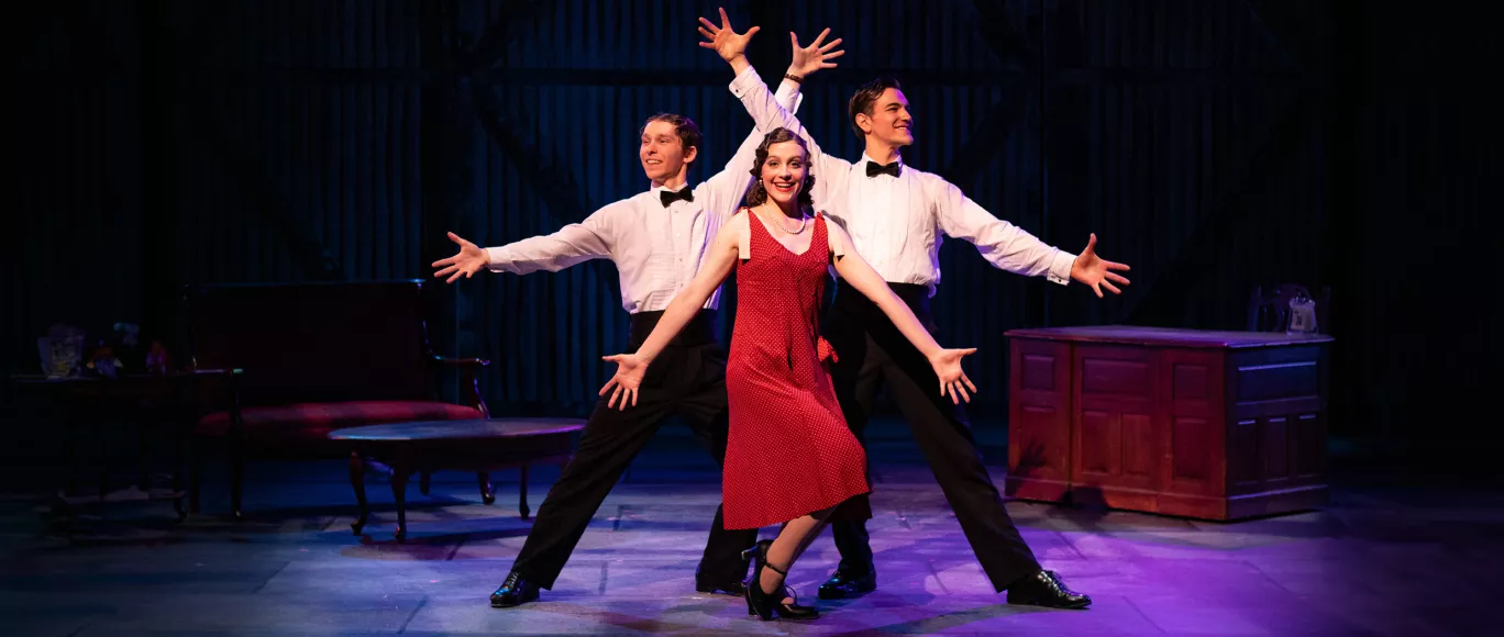 A performer in a red dress outstretches their arms and bevels their leg in front of two other performers in white shirts, bow ties, and black pants. The two performers standing behind outstretch their arms above and beside the performer in front. All performers smile while posing in front of a couch and desk.
