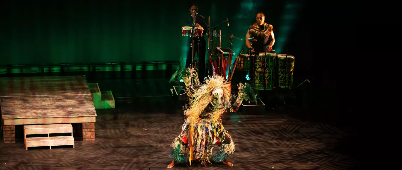 StepAfrika! performer in elaborate costume dances on stage with musicians playing drums in the background.