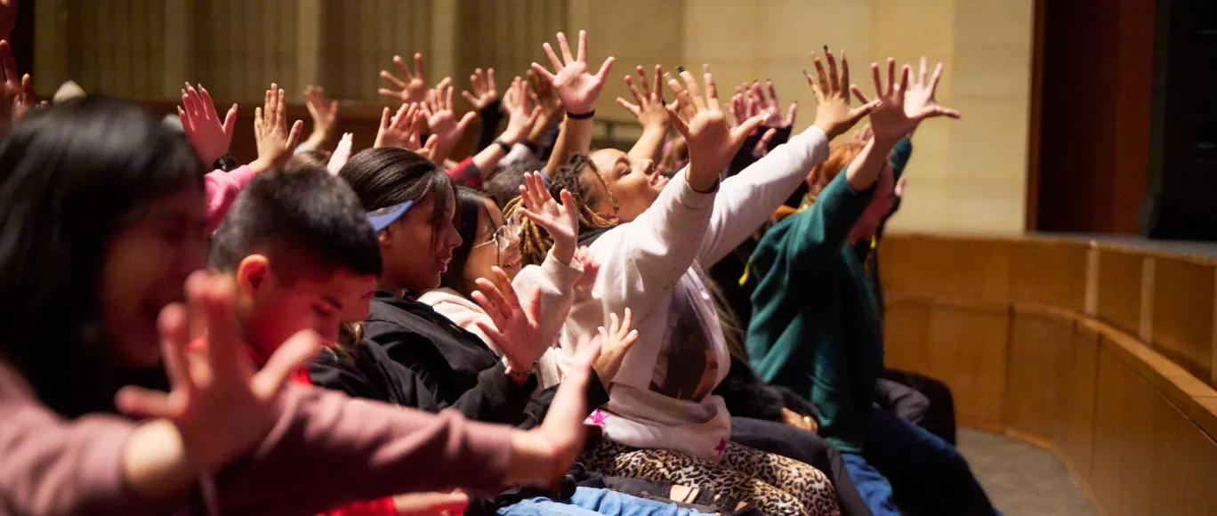 K-12 students wave their hands and reach forward in unison during a performance.