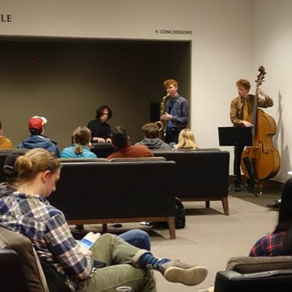 Students gather around musicians playing in the corner of lounge.