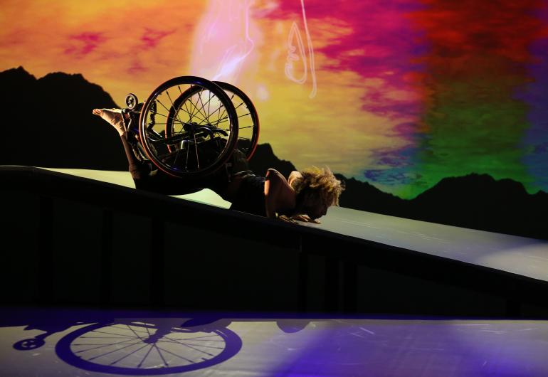 As the sky transitions from gold to amber to red, green and blue, Alice Sheppard, a light-skinned Black woman, slides in a spider position on her stomach down the shiny ramp. The shadow of her wheelchair is visible beneath; her curly hair glows. Photo credit: MANCC / Chris Cameron.