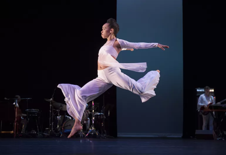 A female dancer wearing white leaps forward on stage.