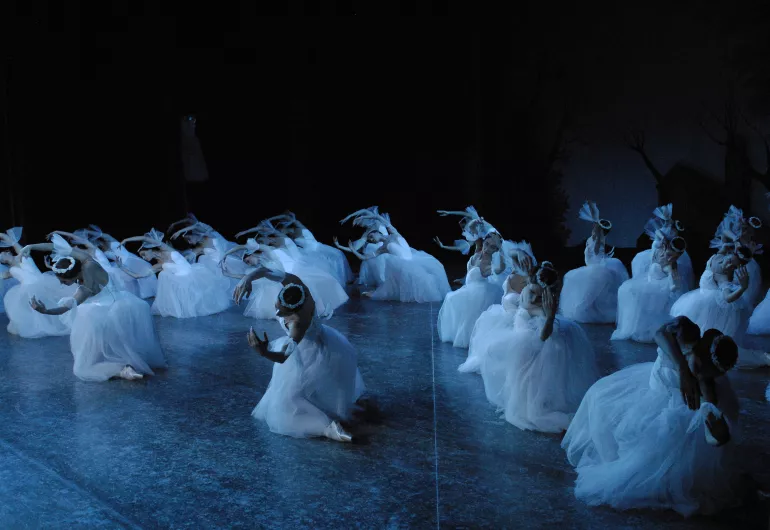 Large group of female dancers in white under a blue light, each on one knee, leaning to the side with rounded arms framing their heads