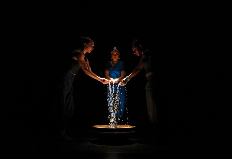 Three dancers in darkness stand around a circle of light letting white grainy substance fall from their fingers into the light.