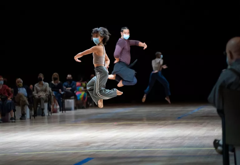 2 dancers wearing face masks jump, legs bent underneath them, arms curved in front