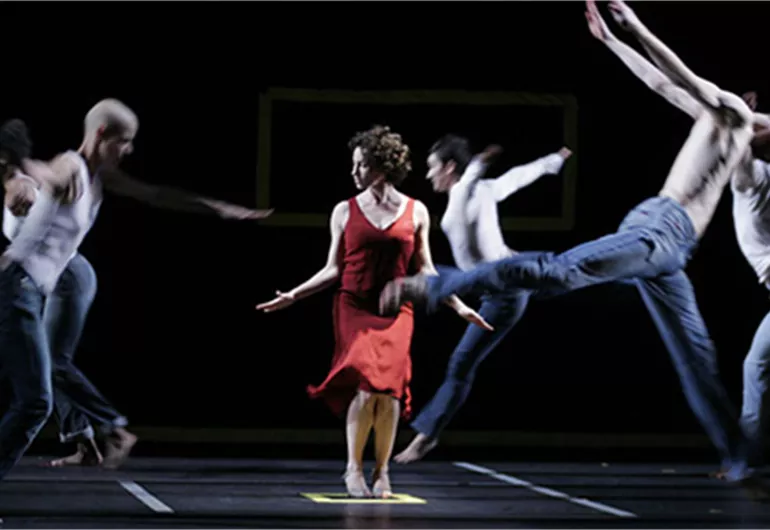 Dancer in red dress posed in the middle of dancers in jeans leaping around her