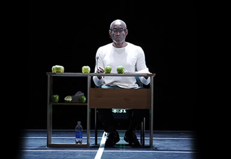 Bill T Jones sits at a desk where a microphone, papers and several apples are placed.