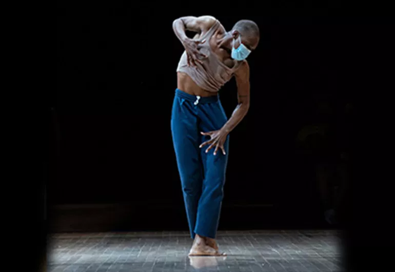 single dancer wearing blue pants and tan tank top and blue facemask, stands in fifth position, hands spread wide on leg and chest
