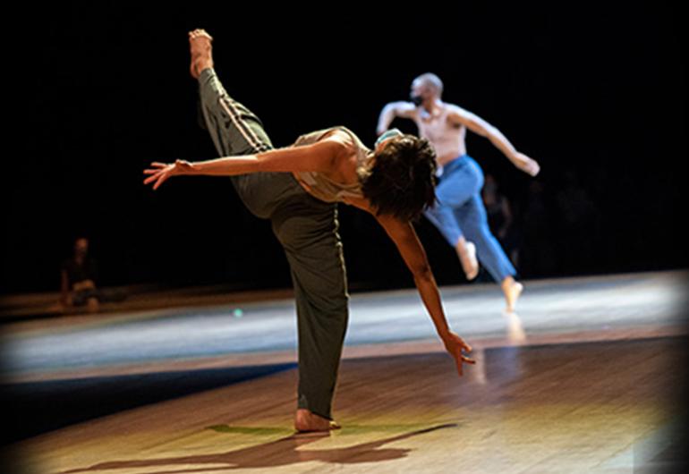 View from behind, one dancer stands on one leg leaning back arms open wide with other leg extended up into the air. Second dancer in background in mid-skip.