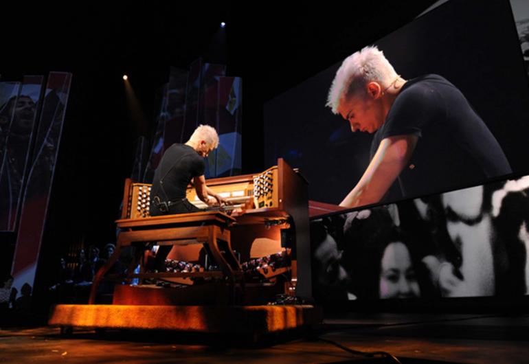 Carpenter plays the organ while a screen displays a closer shot of his profile