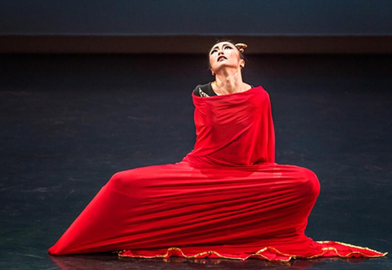 Dancer in stretched red dress in a squating pose looking up with arms behind and one leg out to side
