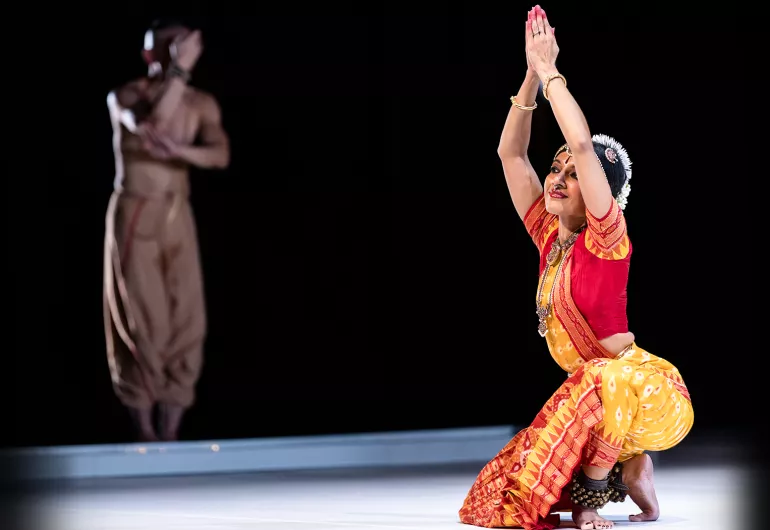 Ragamala dancer is sitting on one leg with arms above her head and palms together