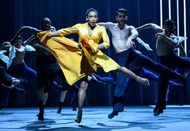 A dancer in a yellow dress strikes a one-legged pose with her remaining arms and legs extended. Other male dancers are in a similar pose behind her. 