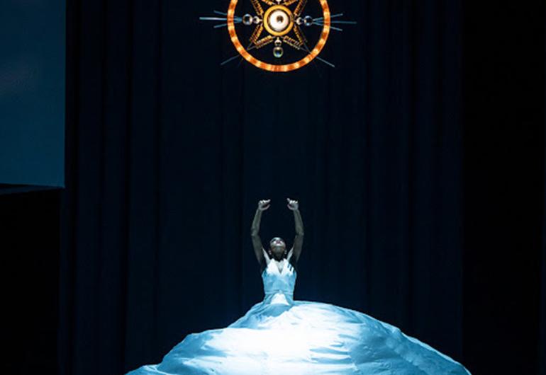 A dancer stands with her arms raised wearing a long white dress that is flared out to create a dome shape.